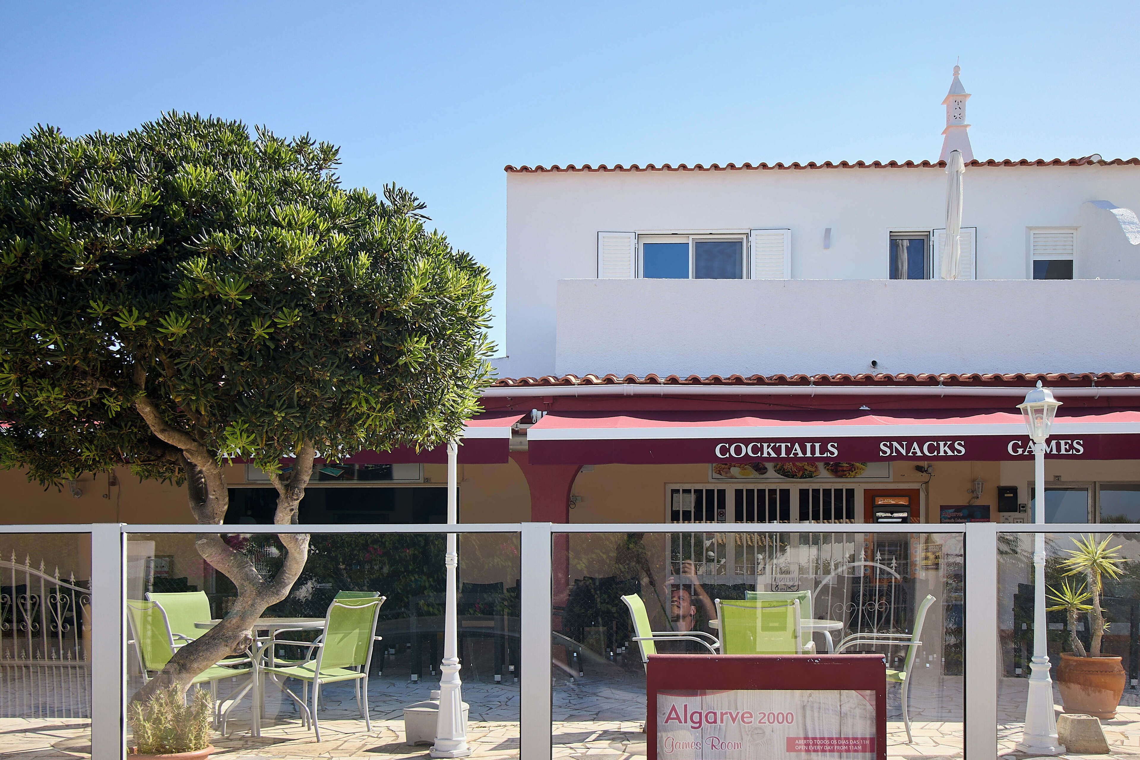 An outdoor seating area with green chairs and a table, a sign for 'Algarve 2000 Games Room', and a building with a tiled roof and a signboard advertising cocktails, snacks, and games.