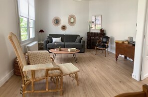 A living room with a grey sofa, two wicker chairs, a round coffee table, a fireplace, and a wooden side table with books. - Léonie's house - 2 bedrooms - private courtyard - wifi (Châteauroux)