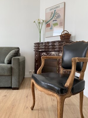 A room with a grey sofa, a wooden chair with a black seat, a wooden fireplace, and a framed map on the wall. - Léonie's house - 2 bedrooms - private courtyard - wifi (Châteauroux)