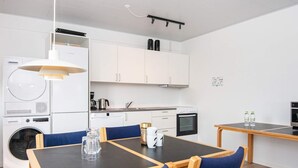 A modern kitchen with white cabinets, a built-in oven, and a coffee machine. There is a dining table with blue chairs and a small table with two bottles on it. - 2 Person Holiday Park Home in Ulfborg (Ulfborg)