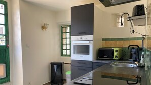 A compact kitchen with a built-in oven, microwave, and toaster. There is a sink with a faucet, a trash bin, and a window with green shutters.