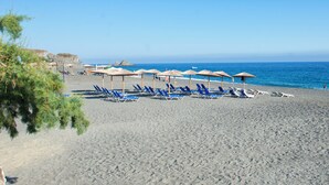 Ein Strand mit blauen Liegestühlen und Sonnenschirmen, ein felsiger Felsen im Hintergrund und das Meer.