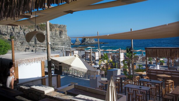 Ein Restaurant direkt am Strand mit Außenbereich, Blick aufs Meer und einem felsigen Felsen im Hintergrund.