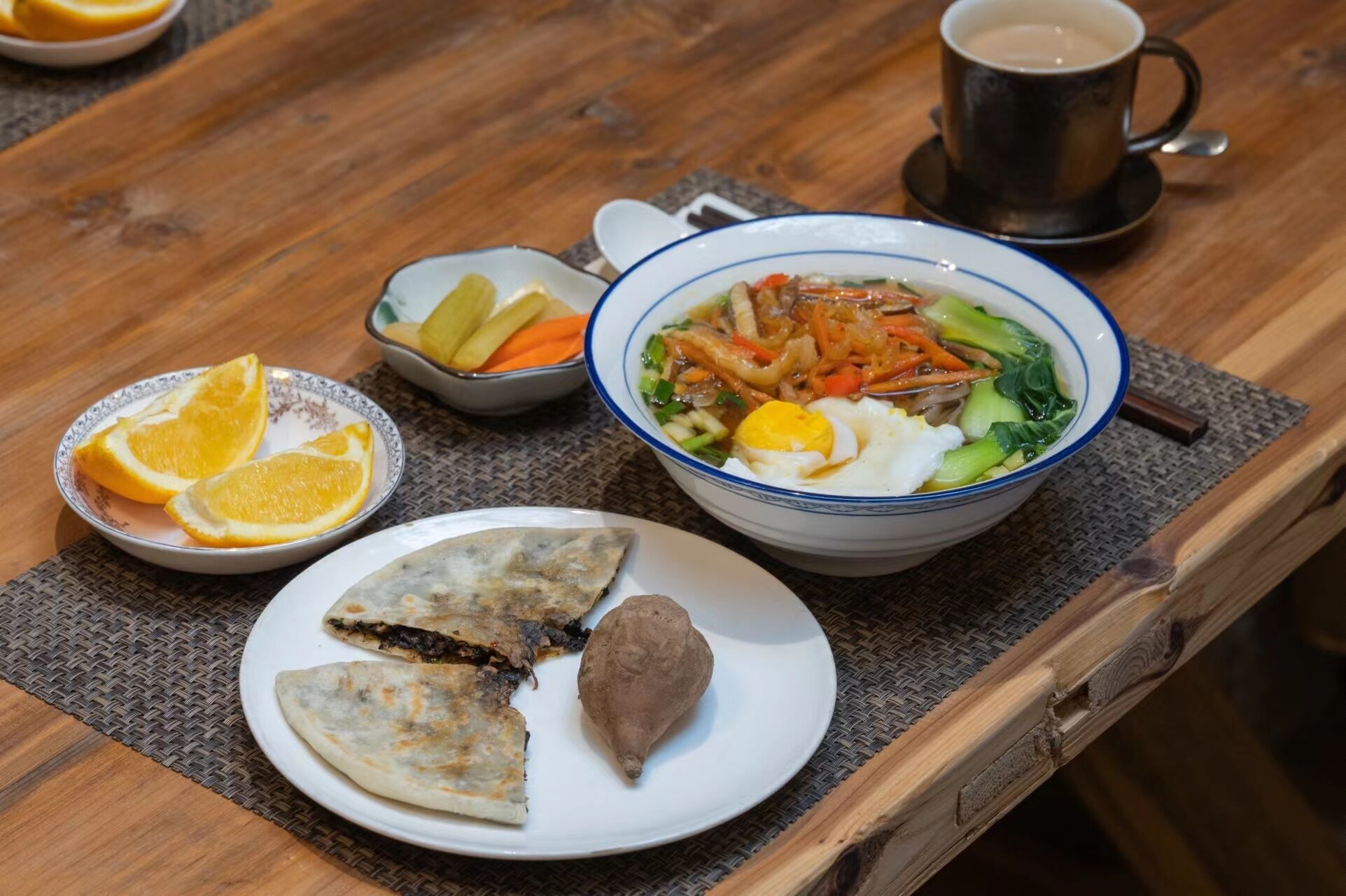 A meal with a bowl of soup, a plate with dumplings, a plate with a fruit, and a cup of coffee on a wooden table.