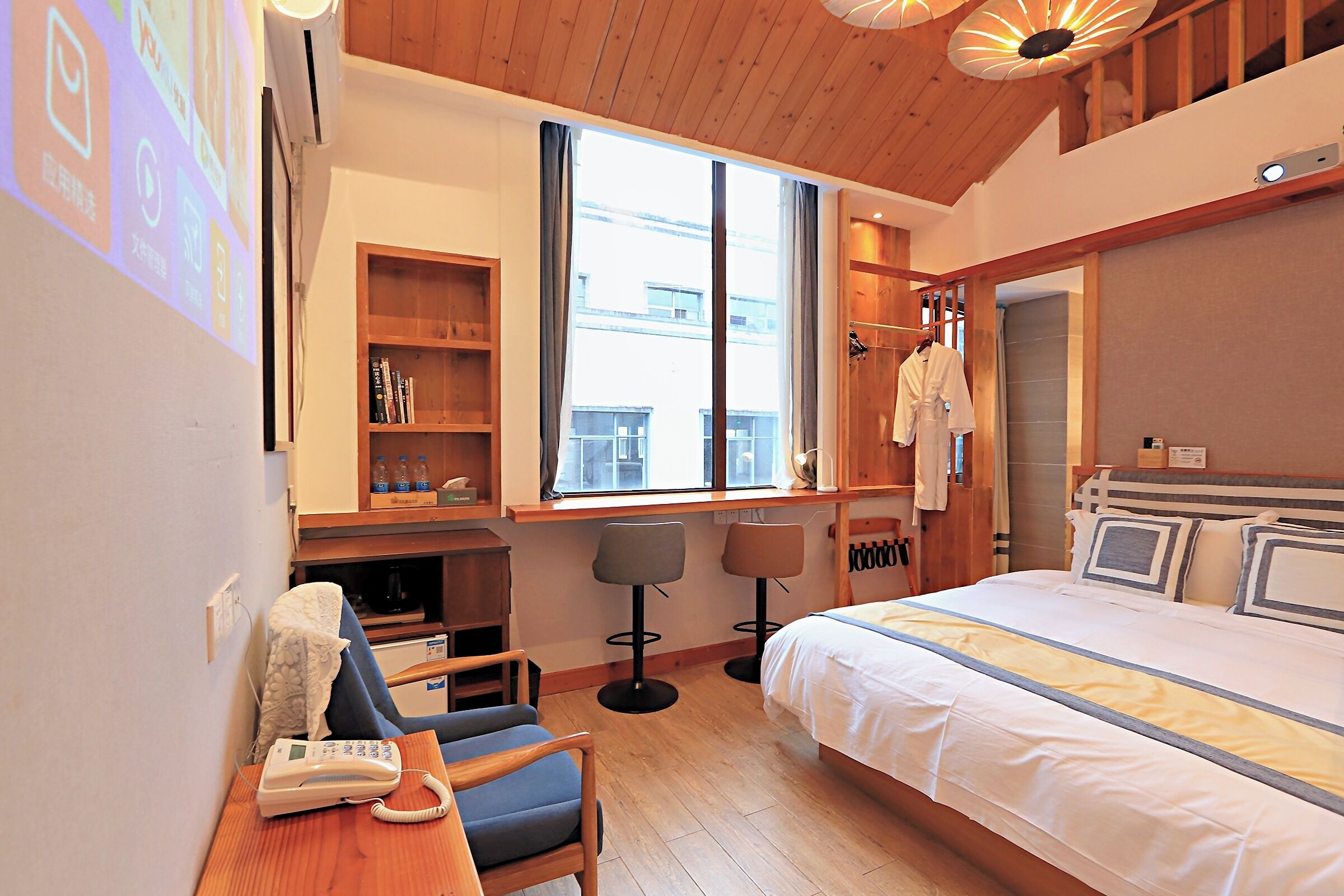 A hotel room with a bed, a desk with a phone, two chairs, a bookshelf, and a window with a view of buildings.