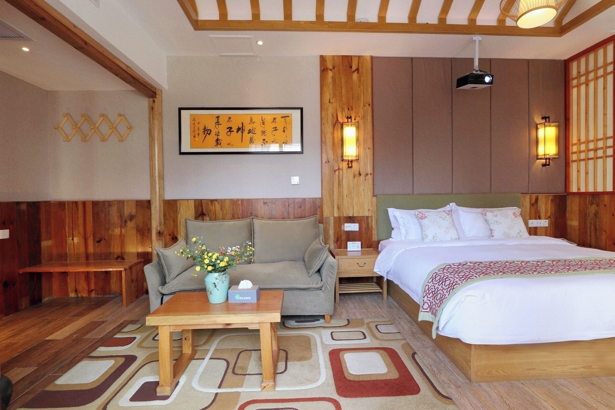 A hotel room with a wooden floor, a sofa, a table with a vase of flowers, a bed with a patterned bedspread, and wall-mounted lights.