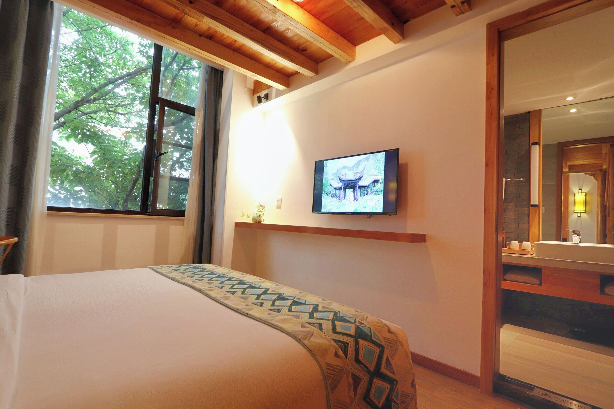 A hotel room with a bed, a television mounted on the wall, a window with a view of trees, and a wooden ceiling.