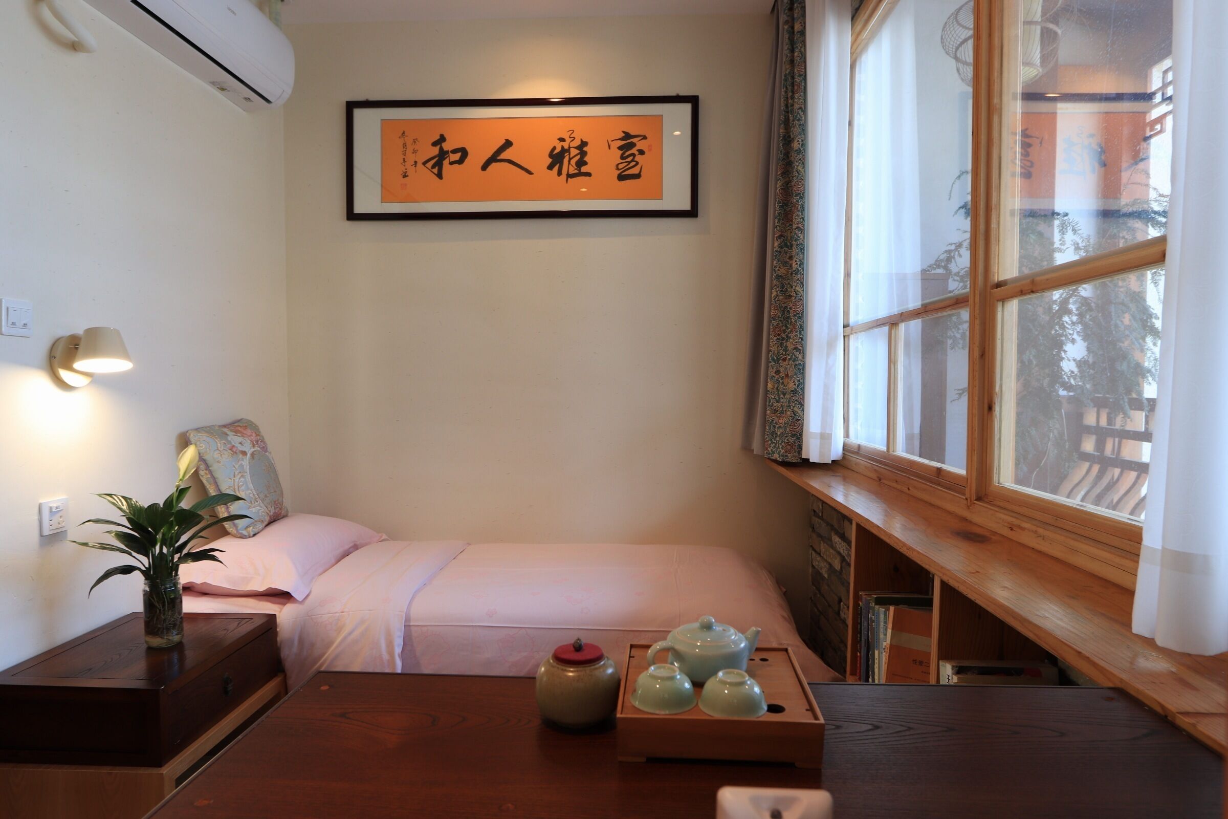 Quarto com cama, mesa de cabeceira com uma planta, bandeja com bules e um quadro na parede.