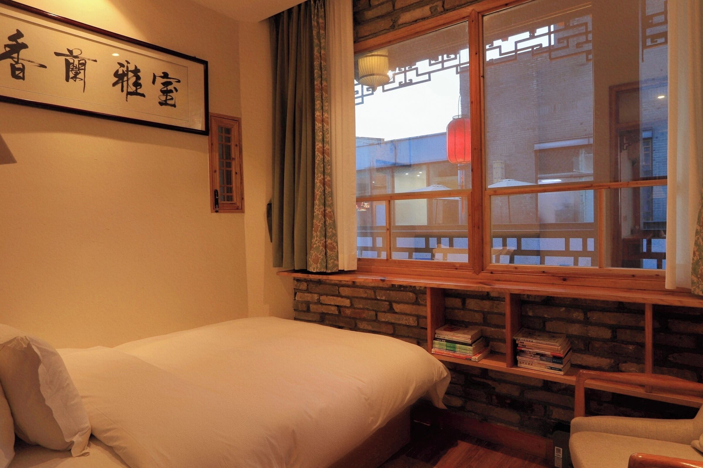 A room with a bed, a bookshelf, and a window with a view of buildings outside.
