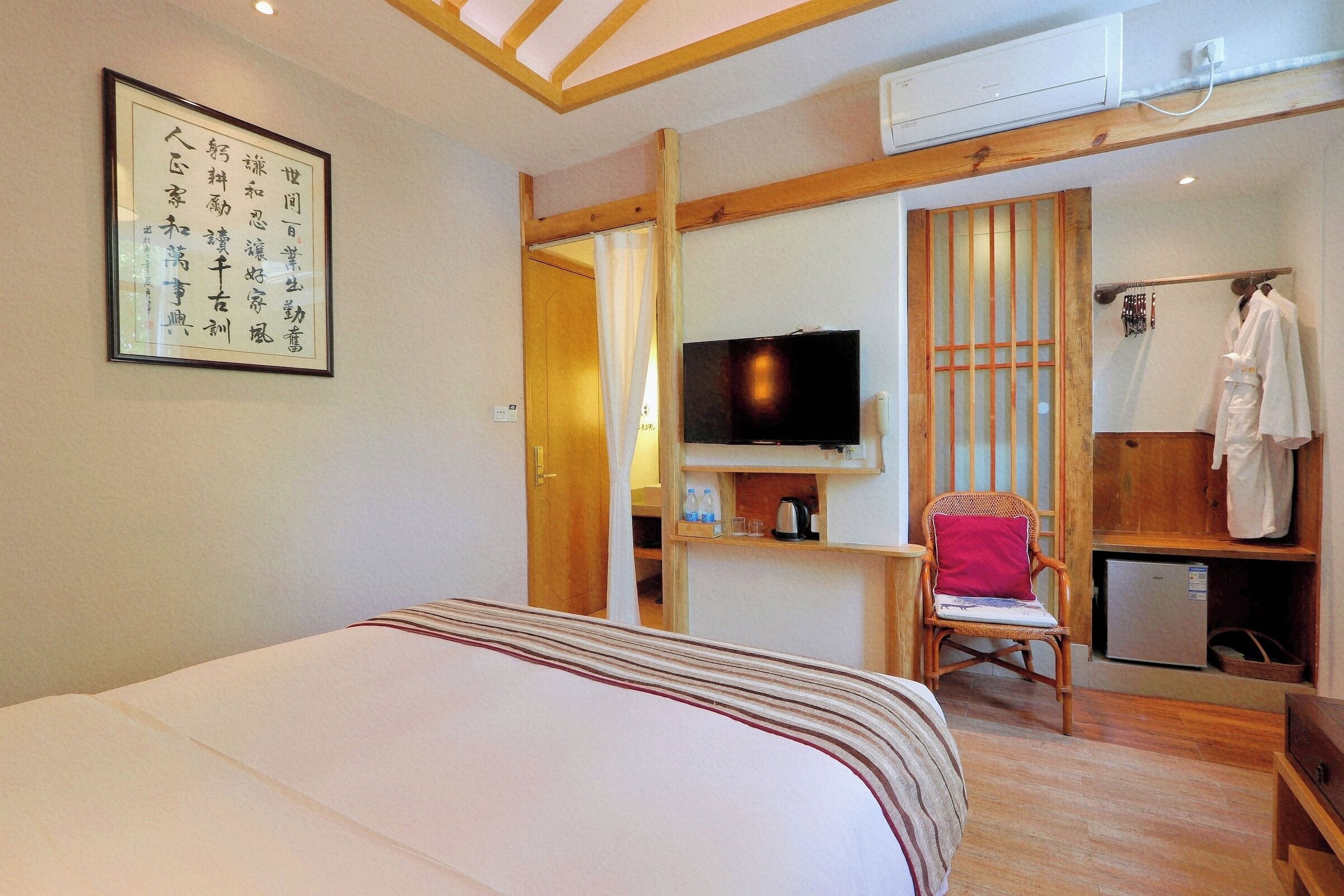 A bedroom with a bed, a television, a wooden chair, a small refrigerator, and a shelf with toiletries.