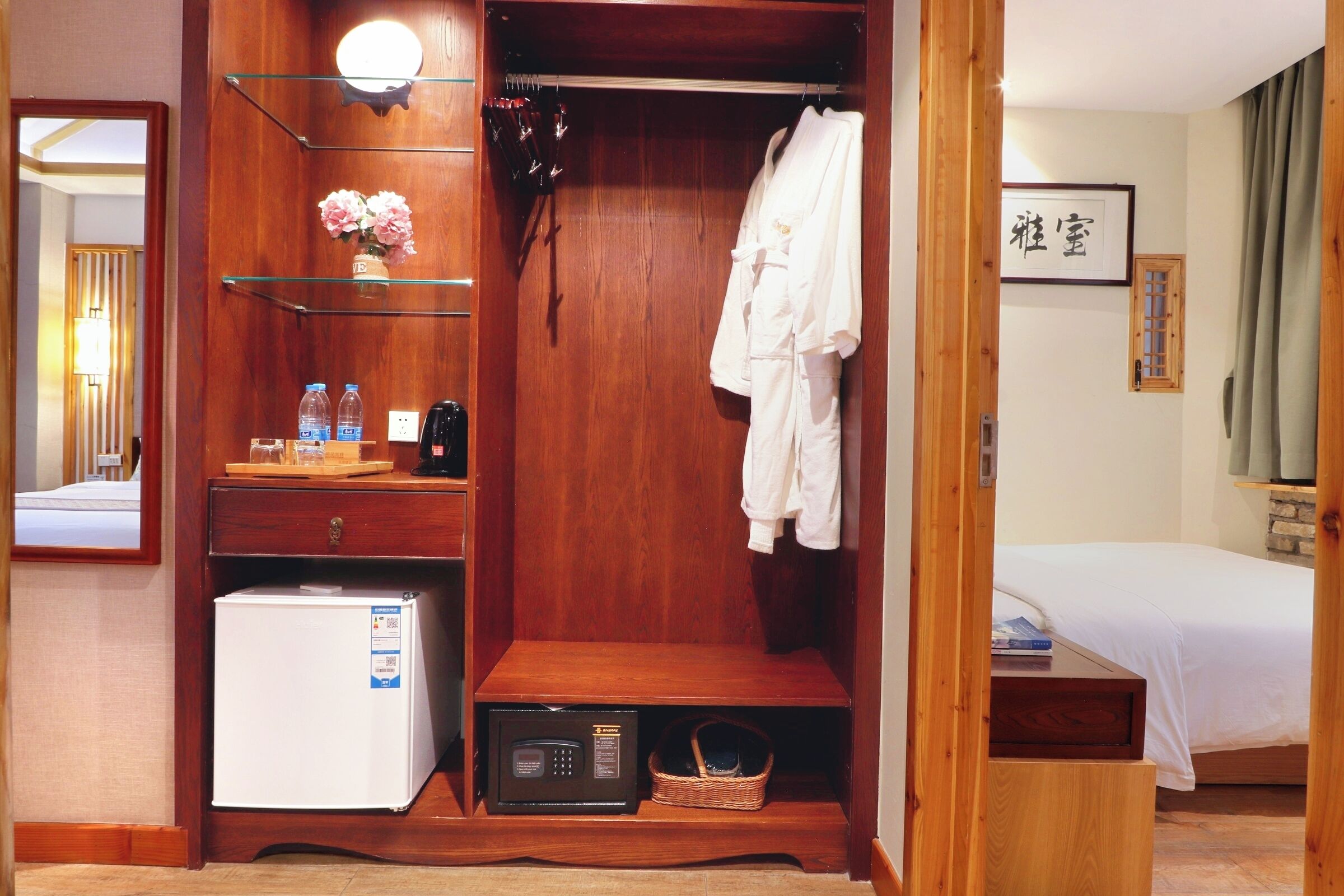 A hotel room with a wooden wardrobe, a bed, a small refrigerator, a microwave, and a shelf with toiletries.