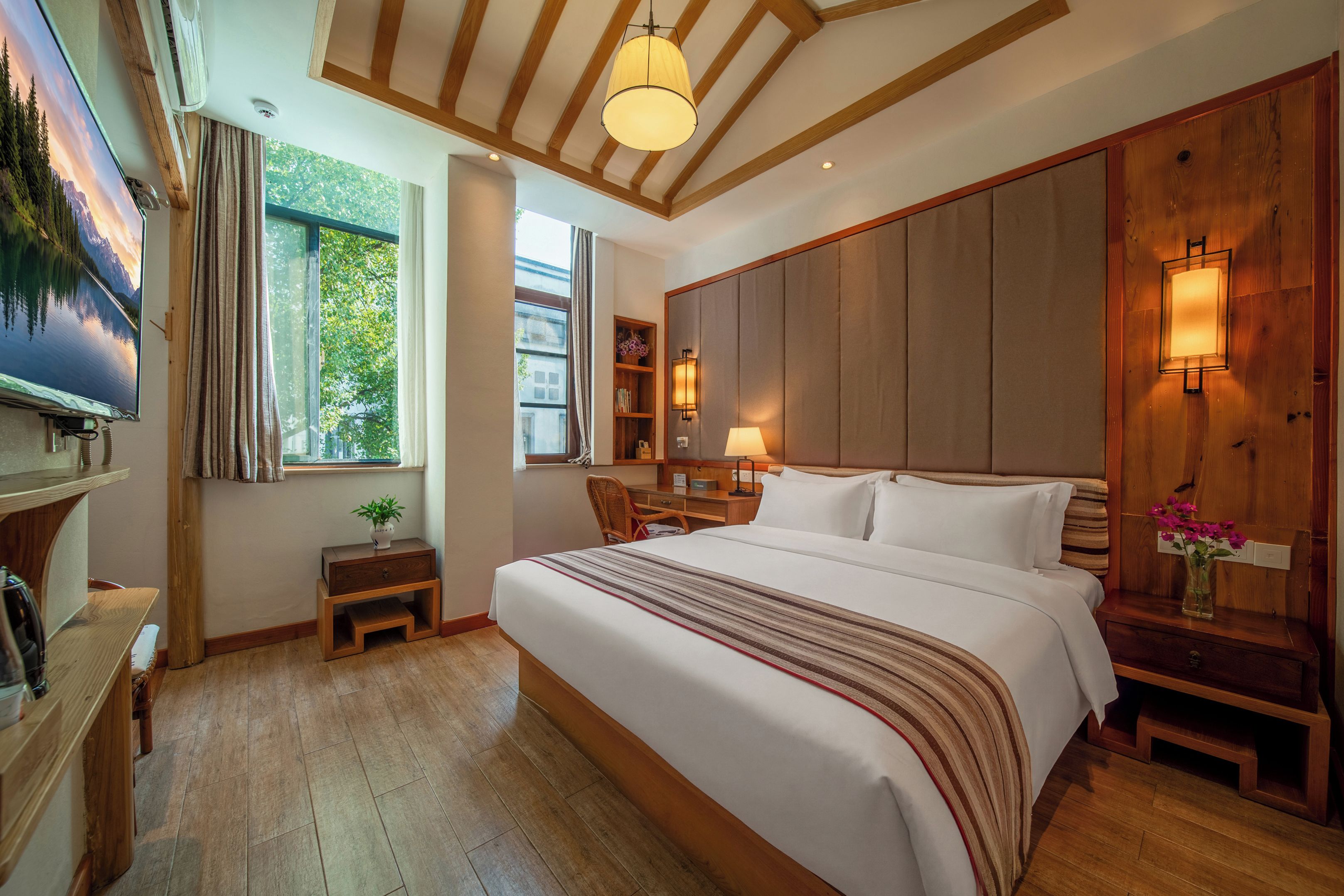 A hotel room with a large bed, a desk, a chair, a nightstand, a lamp, a TV, and a window with curtains.
