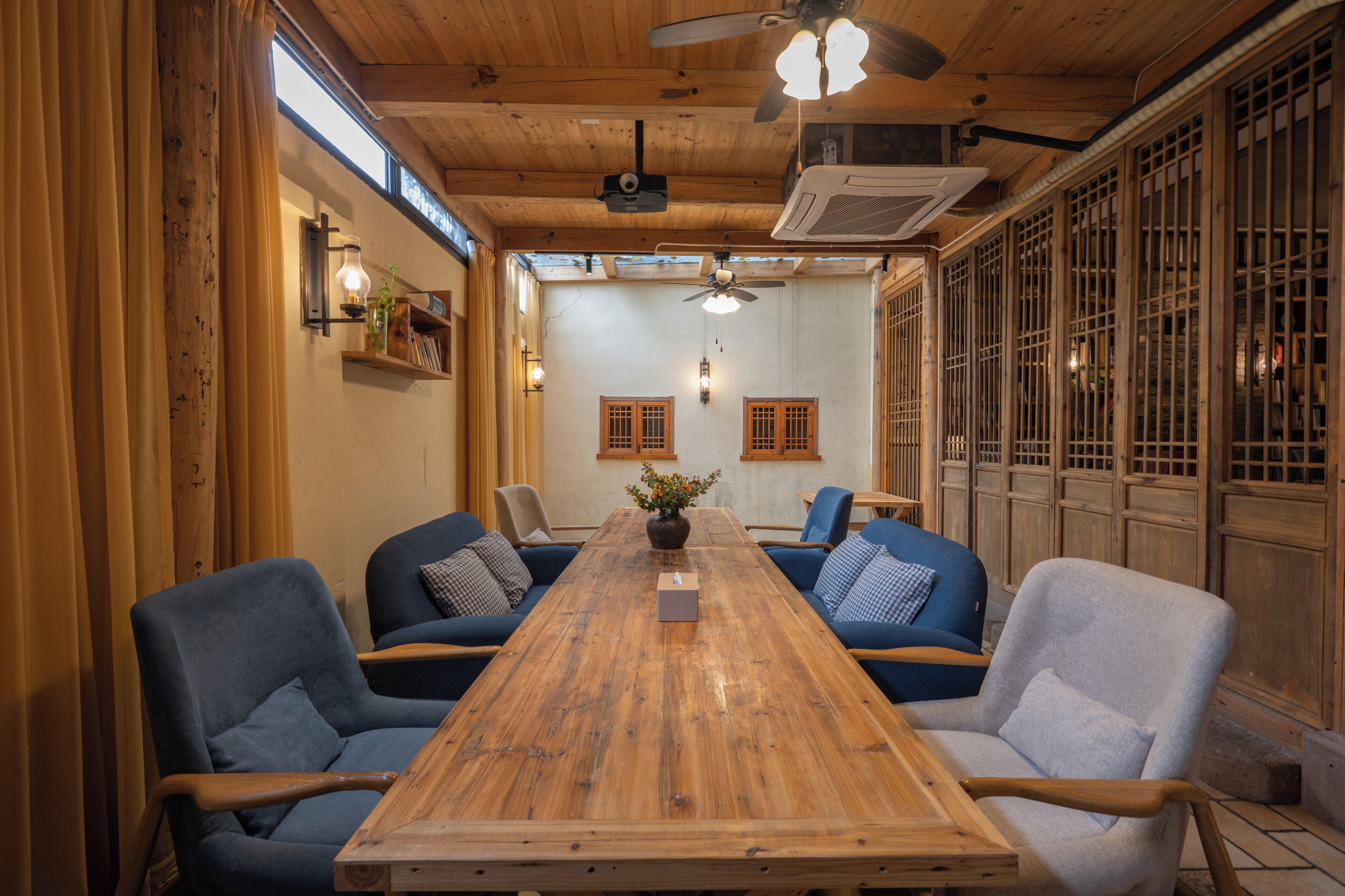 Un salón de conferencias con una mesa de madera, sillas azules y grises, un ventilador de techo y puertas de madera tradicionales.