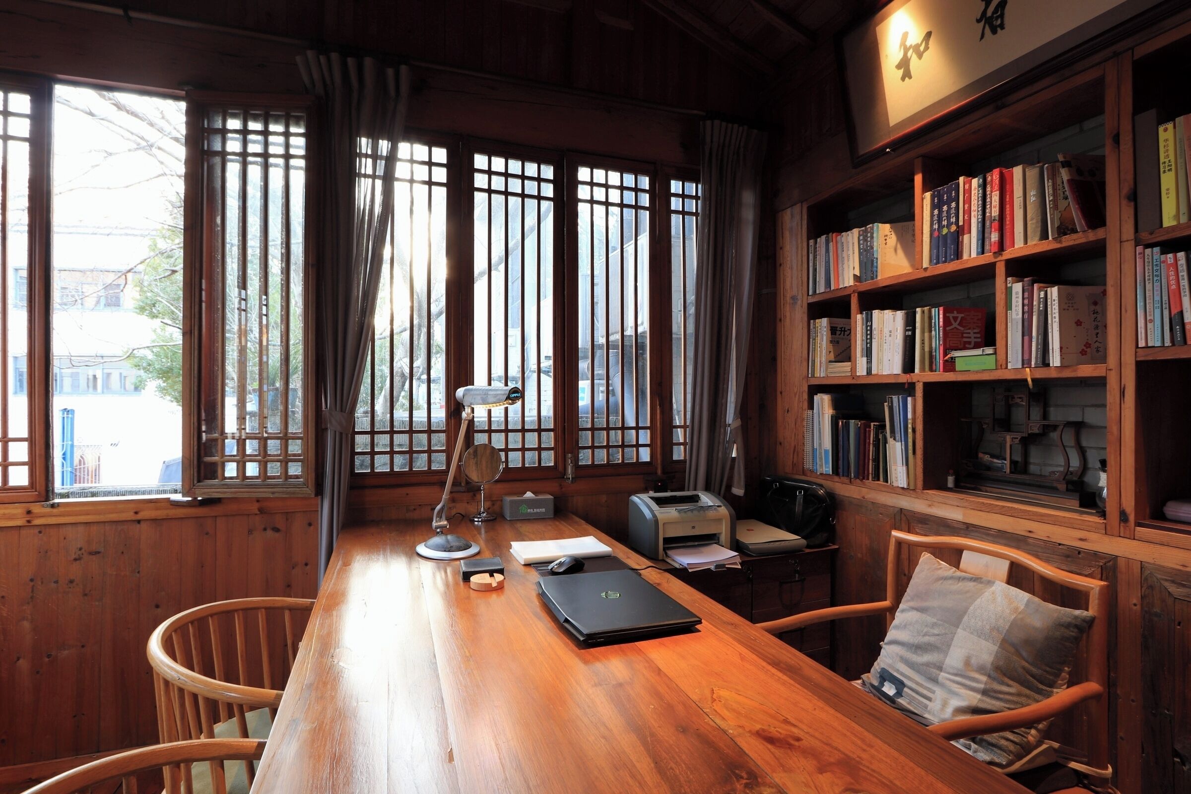 Escritorio de madera con una laptop, una lámpara e impresora. Se ven estanterías llenas de libros y una ventana con cortinas.