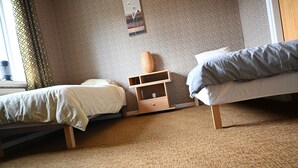 A room with two beds, a small wooden nightstand, a wall-mounted shelf, and a window with curtains.