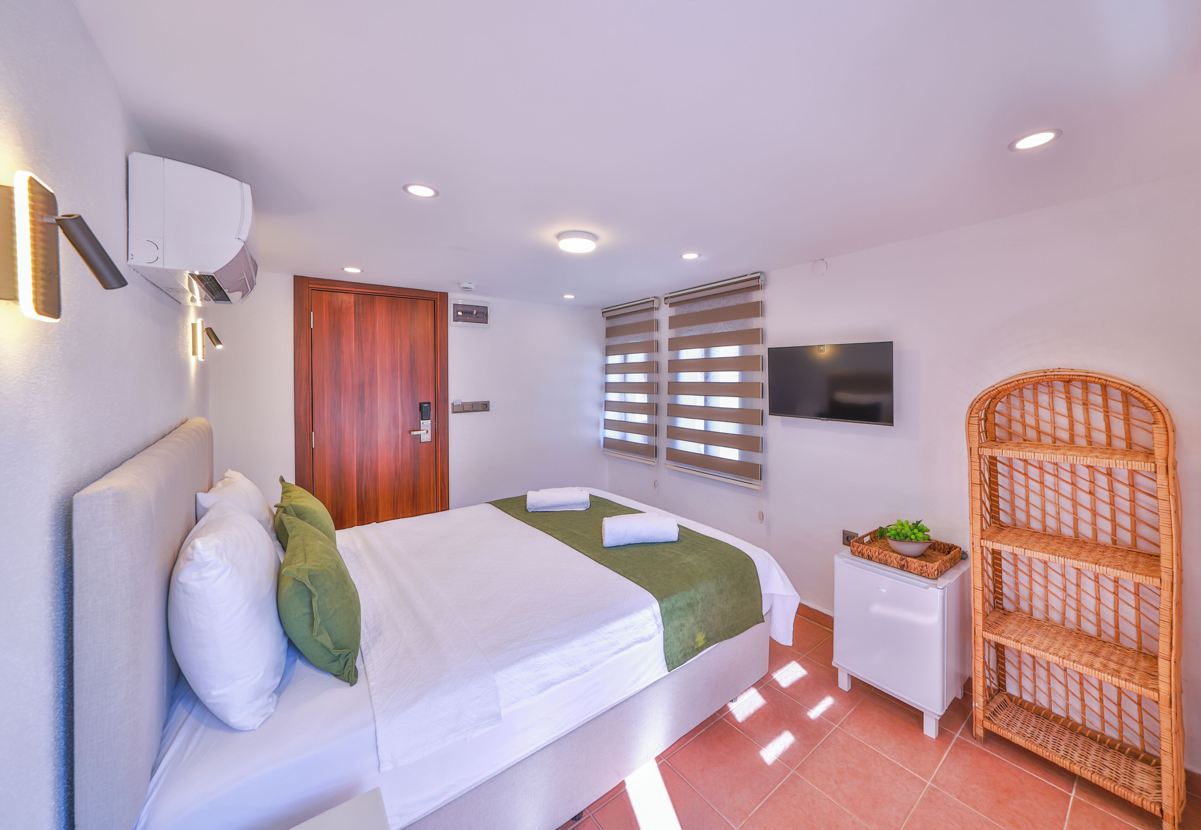 Une chambre d’hôtel avec un lit, une petite table de chevet blanche, une étagère en osier, une porte et une fenêtre avec des stores.