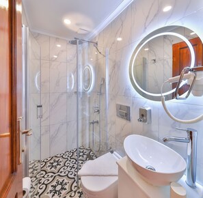 A modern bathroom with a glass shower enclosure, a vanity with a large mirror, and a toilet with a patterned floor. - Kaşhan Hotel (Kas)