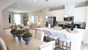 Une cuisine moderne avec des meubles blancs, un îlot central doté de tabourets, une table à manger avec des chaises et un coin salon avec un canapé et une lampe.