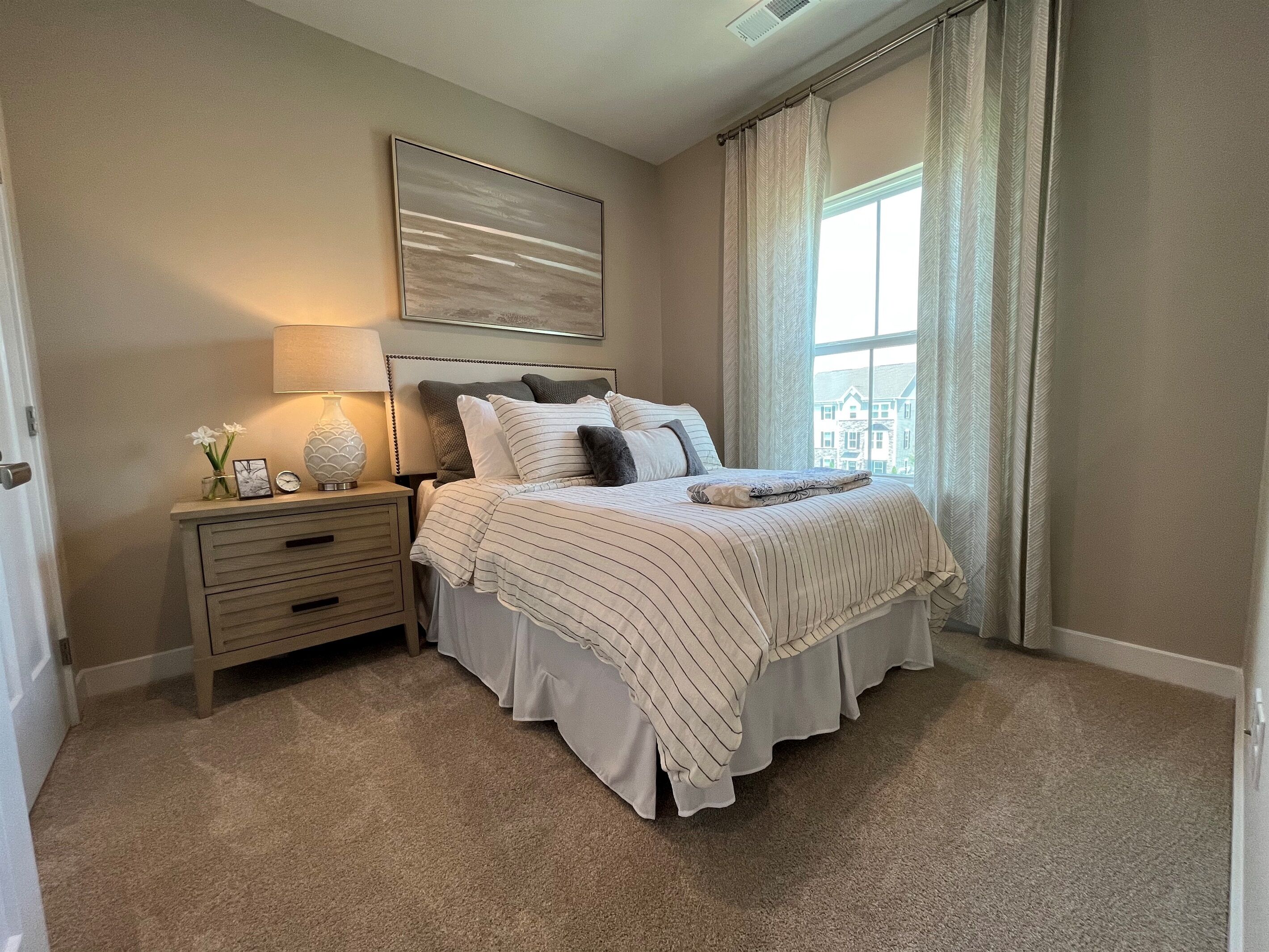 A bedroom with a bed, nightstand, lamp, and a view of a building through the window.