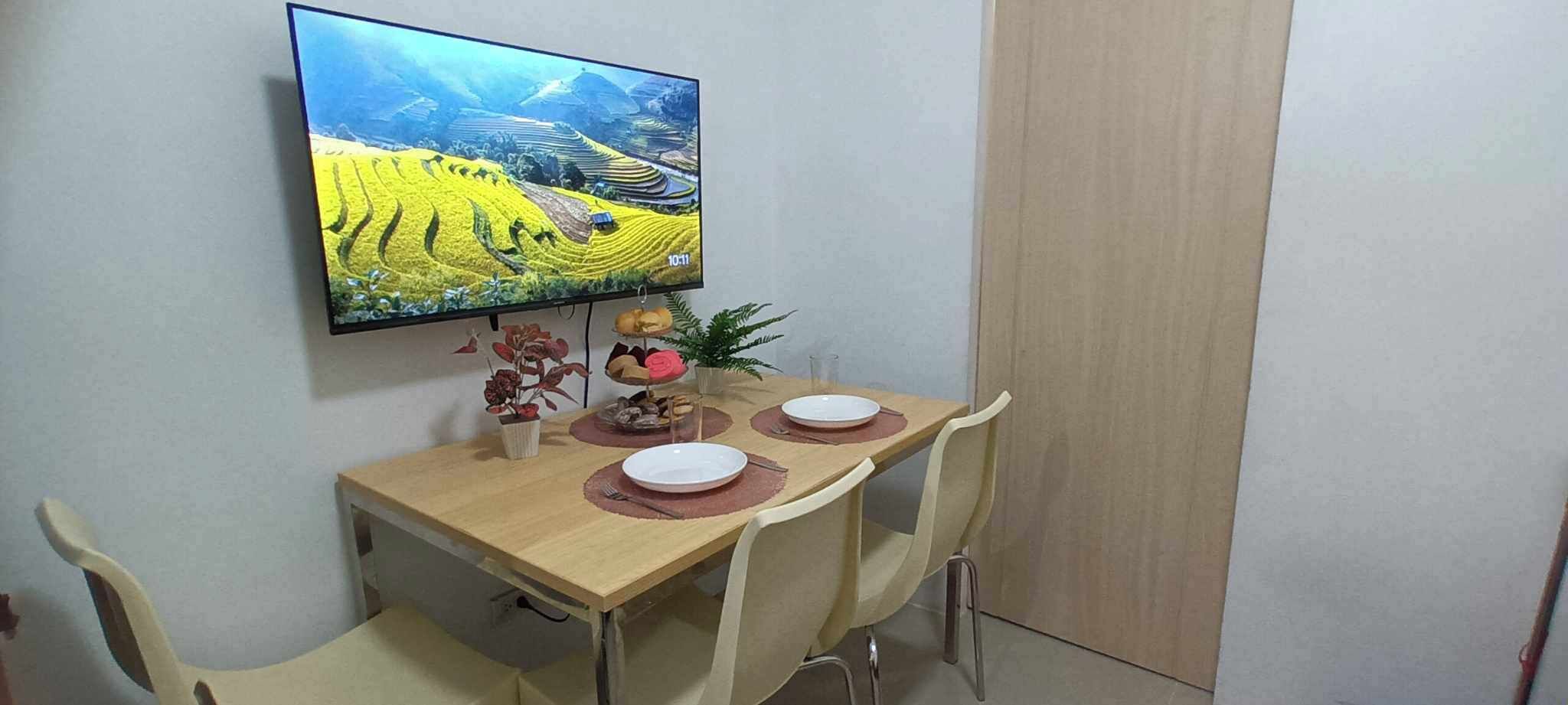 A dining area with a table set for two, a television mounted on the wall, and a door to the right.