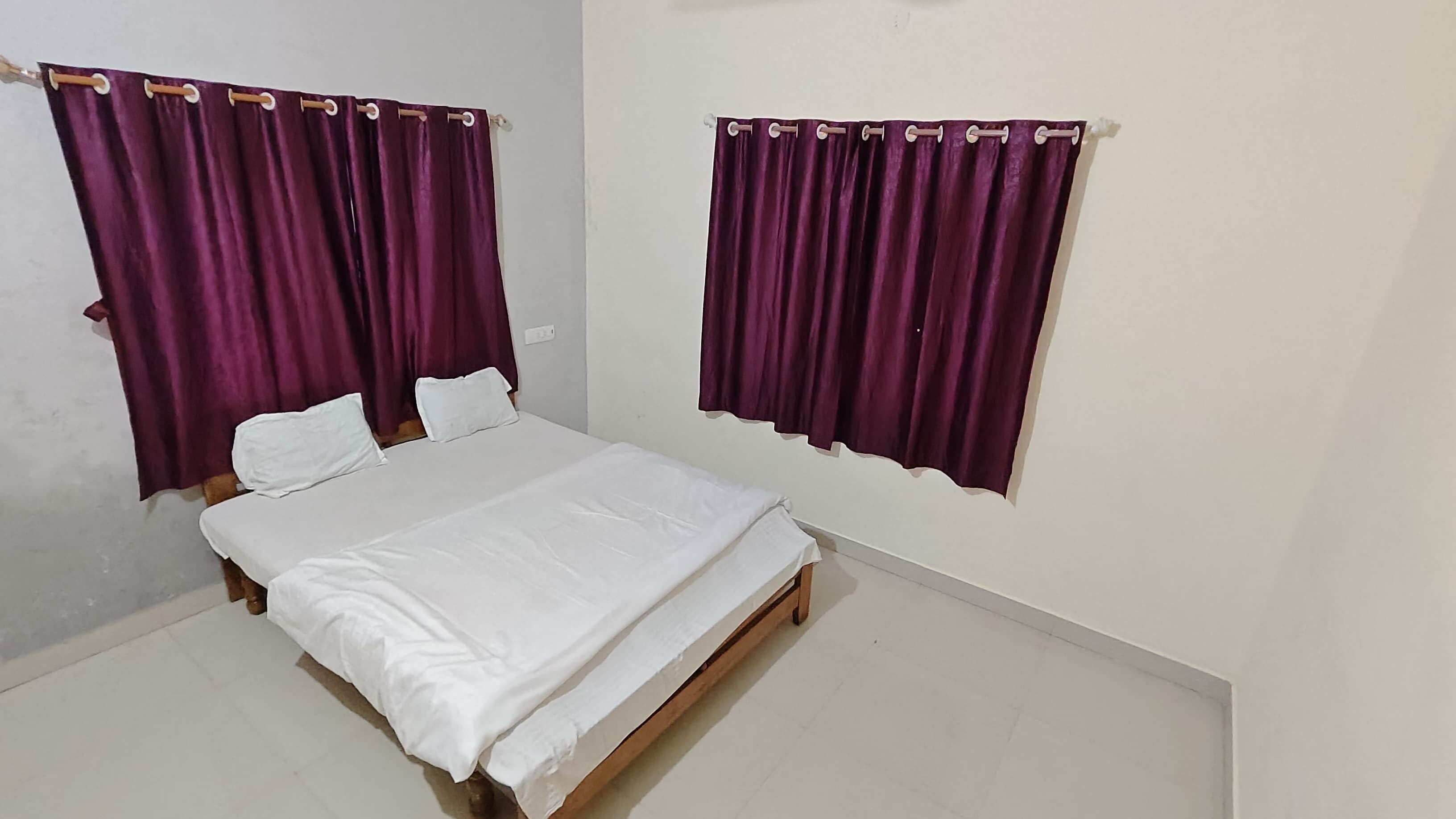 A bedroom with a single bed, two white pillows, and purple curtains on two windows.