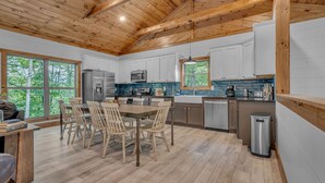A modern kitchen with wooden beams, white cabinets, a dining area with chairs, and a view of trees through large windows. - New!Min Away from Gatinburg,Pool Table, (Gatlinburg)