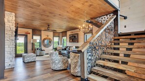 A spacious living area with a wooden staircase, a round mirror, and a view of greenery outside. - Smoky Mtn Luxury Cabin, FREE Waterpark, PS5, Pool Table, Indoor Pool, Mini Golf (Sevierville)