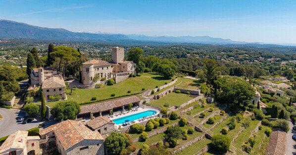 A castle with a swimming pool, surrounded by greenery and a mountainous backdrop.