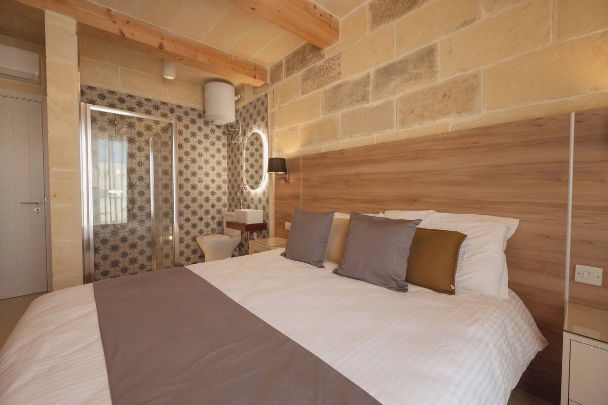Une chambre dotée d’un grand lit, d’une tête de lit en bois, d’un espace douche et d’une salle de bain avec un miroir et un porte-serviettes.