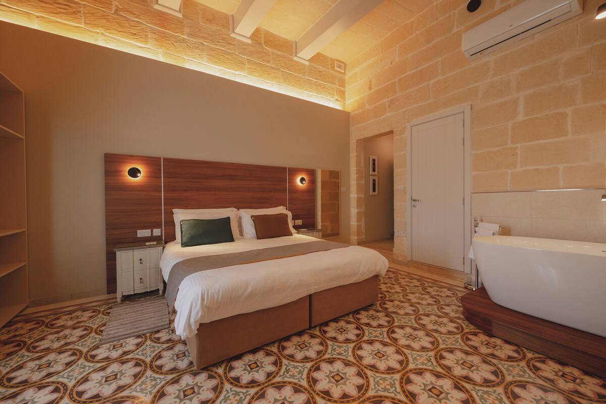 Une chambre à coucher avec un grand lit, une tête de lit en bois, une banquette intégrée, une baignoire blanche et un sol à motifs.
