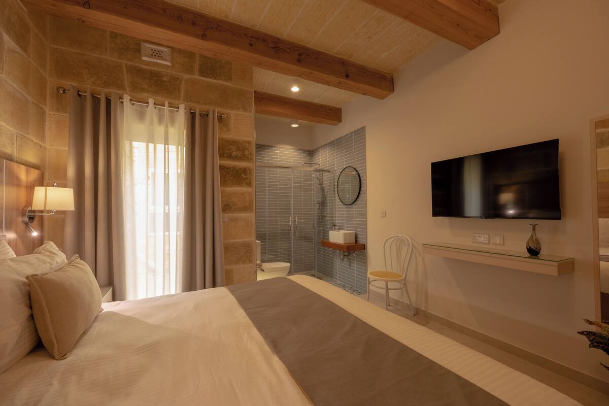 Une chambre avec un grand lit, un téléviseur fixé au mur, une salle de bain avec douche et toilettes, et un plafond en bois.