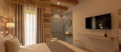 A bedroom with a large bed, a TV mounted on the wall, a bathroom with a shower and toilet, and a wooden ceiling.