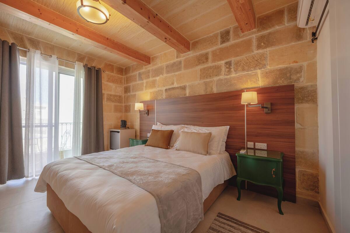 Une chambre avec un grand lit, une tête de lit en bois, deux lampes de chevet et un mur en pierre.