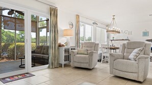 Ein geräumiges Wohnzimmer mit einer Glastür zum Patio, einem bequemen Sofa, zwei Sesseln, einem kleinen Tisch und einer Lampe.