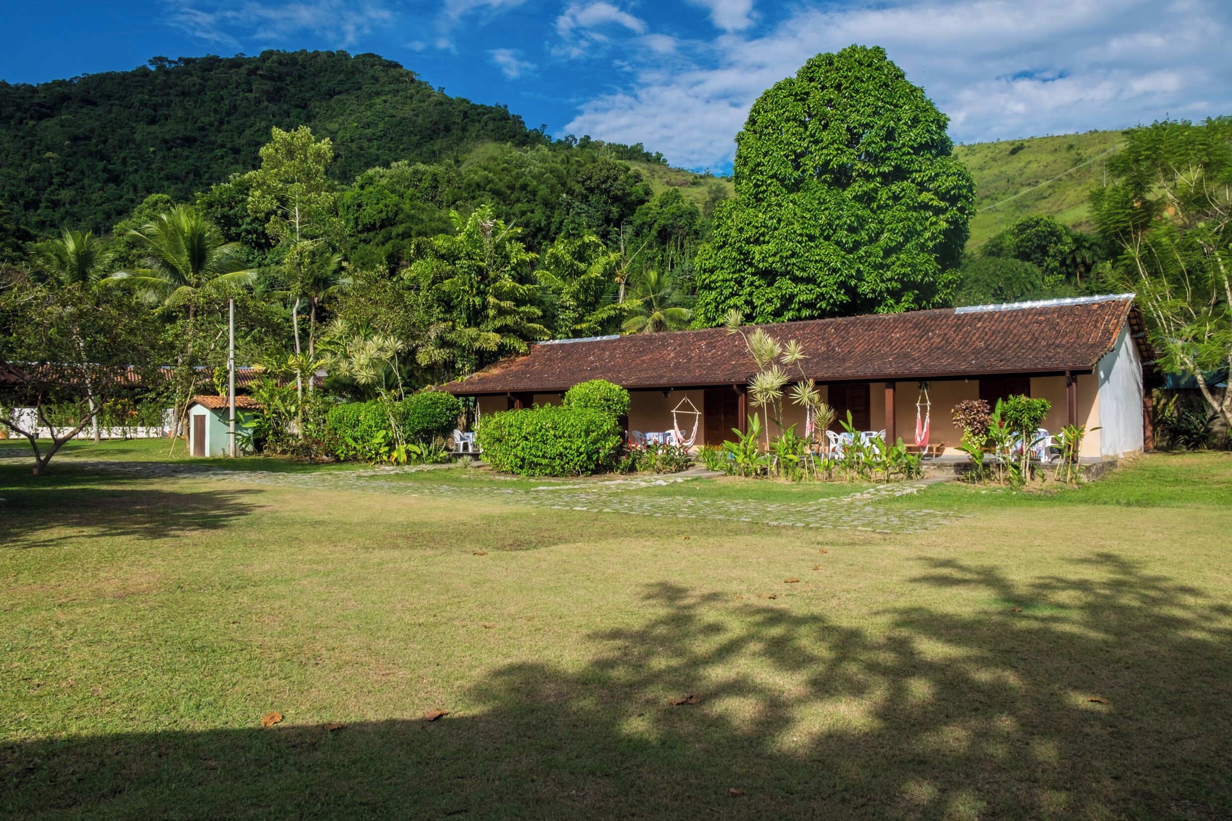 A rustic building with a tiled roof, surrounded by lush greenery and a hill in the background.