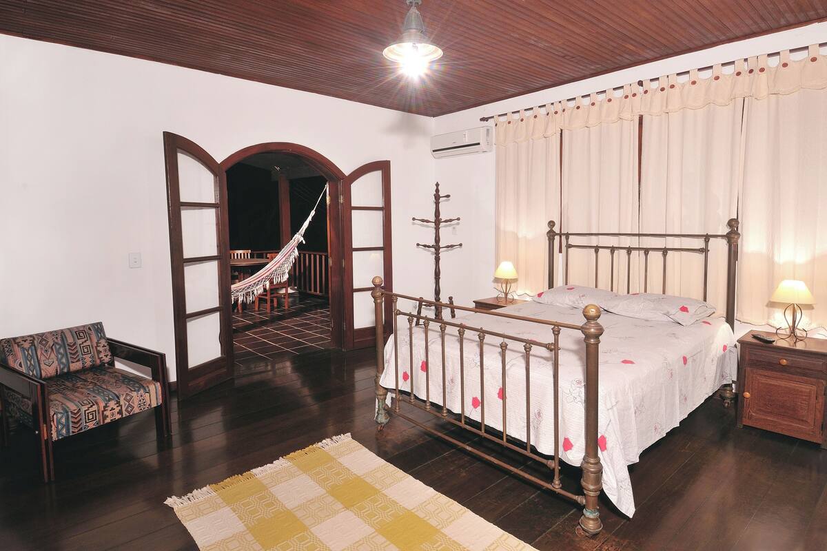 A bedroom with a wooden bed, a nightstand, a chair, a patterned rug, and a hallway leading to another room.
