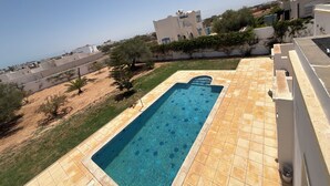 A rectangular swimming pool with a tiled border, surrounded by a grassy area and a few trees. - Villa mayline (Arkou)