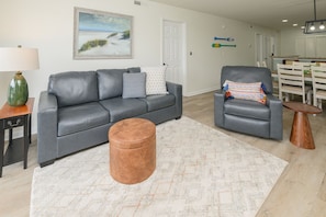 Ein Wohnzimmer mit grauem Sofa, pouf, Teppich mit Muster und einem Essbereich mit weißem Tisch und Stühlen.