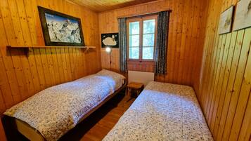 Une chambre de style cabane en bois, avec deux lits simples, une fenêtre avec des rideaux, un tableau accroché au mur et une lampe fixée au mur.
