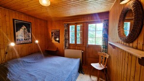 A wooden cabin room with a bed, a chair, a radiator, and a window with curtains.