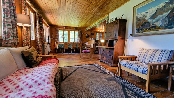 A cozy cabin interior with a wooden ceiling, a sofa, a chair, a dining table, and a fireplace.