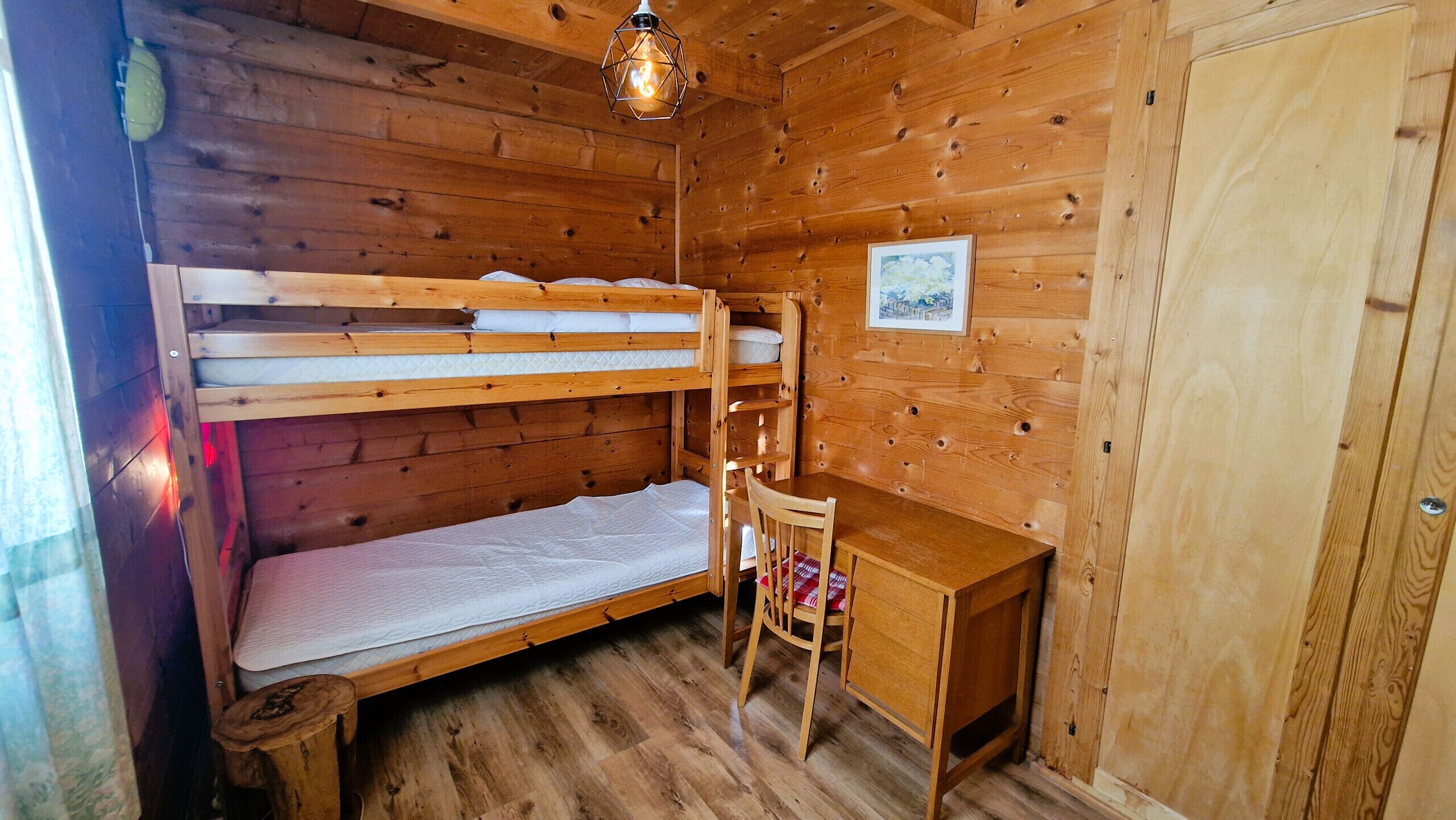 A wooden bunk bed room with a desk and chair, a hanging light fixture, and a framed picture on the wall.
