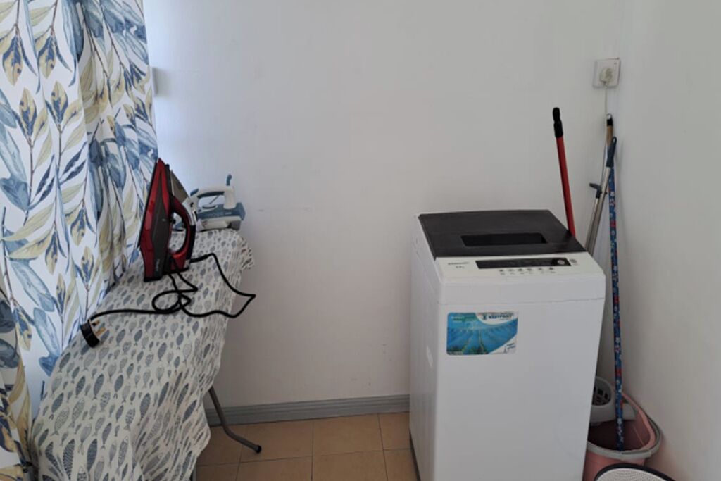 Una habitación con cortina estampada, un planchado de hierro rojo, una lavadora blanca y una mopa apoyada en la pared.