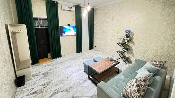 Una sala de estar con un sofá, una mesa de centro y un televisor. Hay una ventana con cortinas, un reloj en la pared y una mesita con un jarrón.
