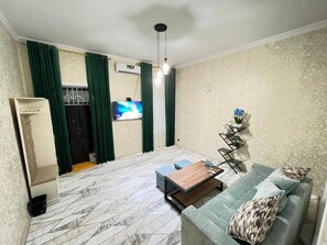 A living room with a sofa, a coffee table, and a TV. There is a window with curtains, a clock on the wall, and a small table with a vase. - All inclusive  (Samarkand)