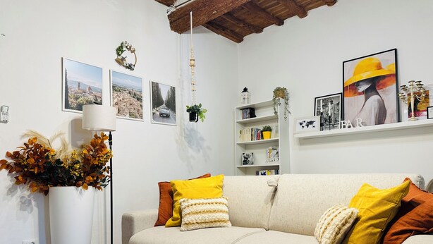 A living room with a beige sofa, yellow and brown cushions, a white side table with a remote control, a floor lamp, and a bookshelf with books and decorative items. - Beautiful air-conditioned apartment a few meters from the center and tram stop. (Firenze)