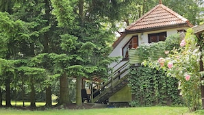 A building with a red-tiled roof, a staircase leading to a balcony, and a garden with trees and flowers. - Cozy vacation apartment in the middle of nature, with terrace and barbecue area (Hinzenhagen)