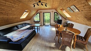 A cozy attic room with a black leather sofa, a wooden dining table with wicker chairs, and a television on a small table. - Cozy vacation apartment in the middle of nature, with terrace and barbecue area (Hinzenhagen)