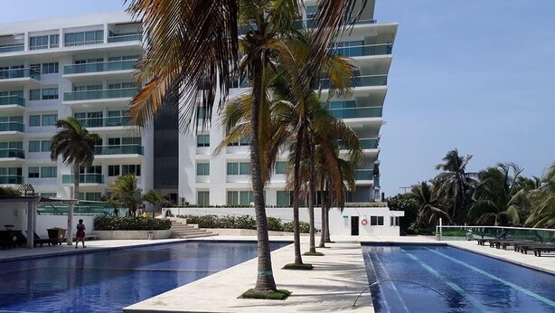 A swimming pool with palm trees, a white building with balconies, and a clear sky. - Luxury Entire 2 BR Condo Oceanfront In Cartagena (Cartagena)