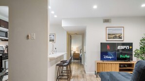 A modern living room with a TV displaying Netflix, Disney+, ESPN+, and Hulu. There is a kitchen area with a stove and a dining area with a bar stool. - East Side #212 xxxxxxx (Austin)